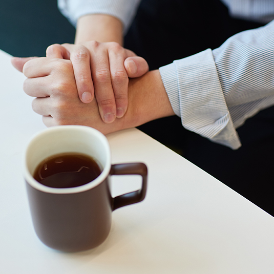 Photo is a close-up of Keny’s crossed hands next to a cup of coffee