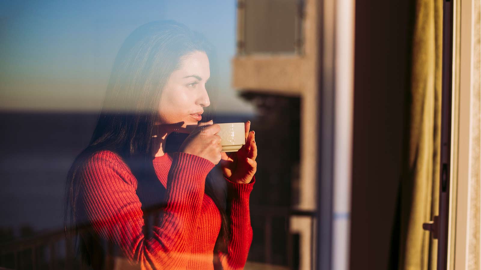 A younger woman drinking from a cup and looking out of the window into the distance.