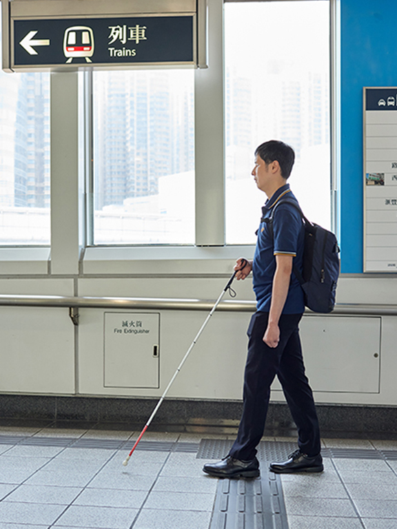 Photo shows Keny in casual clothes walking in the subway with his cane, with a sign above pointing to the trains