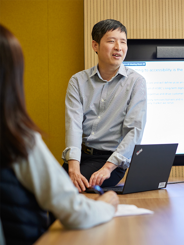 Photo shows Keny in office clothes perched against a desk and leading a presentation to colleagues