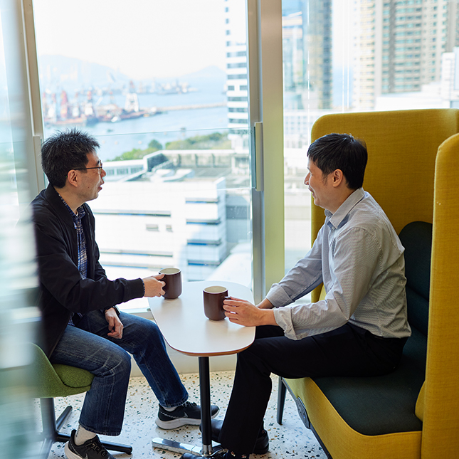 Photo shows Keny and a colleague in conversation with cups of coffee