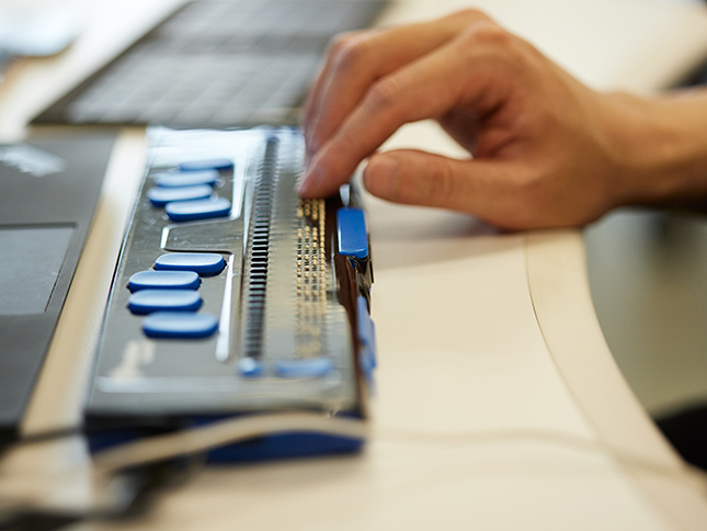 Photo is close-up of Keny’s fingers typing on a Braille keyboard