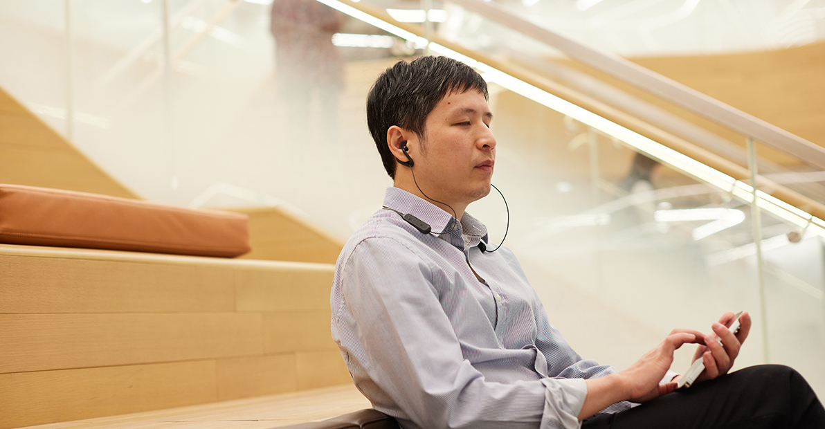 Photo shows Keny in office clothes relaxing in the HSBC office and reading on his phone, with his headphones on