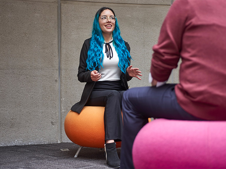 Blue hair woman in black clothes talking with a men in red shirt and jeans, both sitting in colorful stools