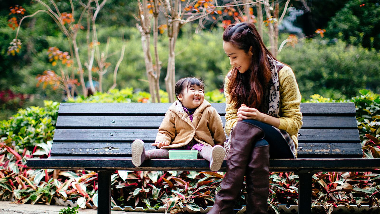 A mother laughs with her young daughter on a park bench