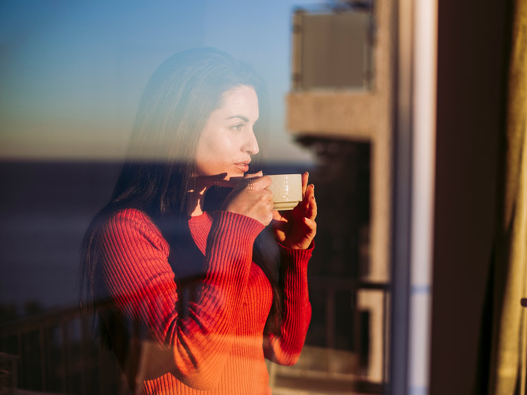 A younger woman drinking from a cup and looking out of the window into the distance.